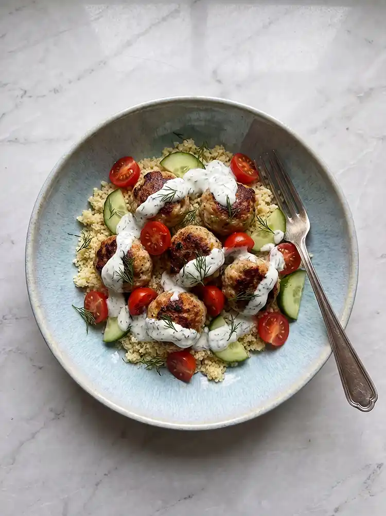 Overhead view of a Mediterranean chicken meatball bowl with grains, tomatoes, cucumber, and tzatziki drizzle