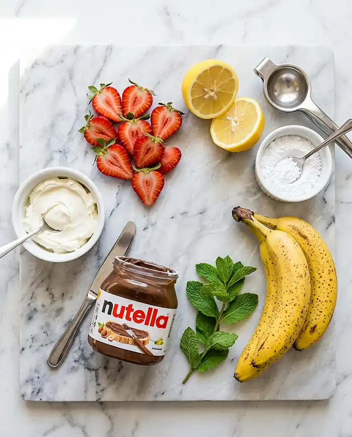 A flat lay of sweet crepe filling ingredients including cream cheese, strawberries, lemon, Nutella, and powdered sugar