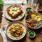Three high protein dinner recipes arranged on a wooden table including egg white wrap lasagna, honey garlic shrimp, and chipotle chicken bowl