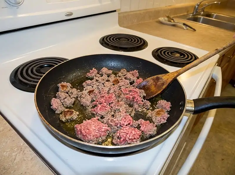 Mini Cottage Pies 4 mince browning in the scratched black nonstick frying pan, part still pink and raw, fat pooling, wooden spoon mid-stir